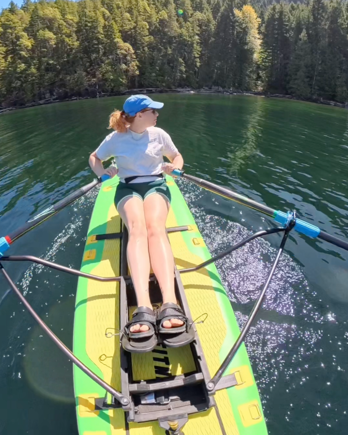 A woman rows on a bright green and yellow paddleboard on a calm lake with a dense forest in the background.