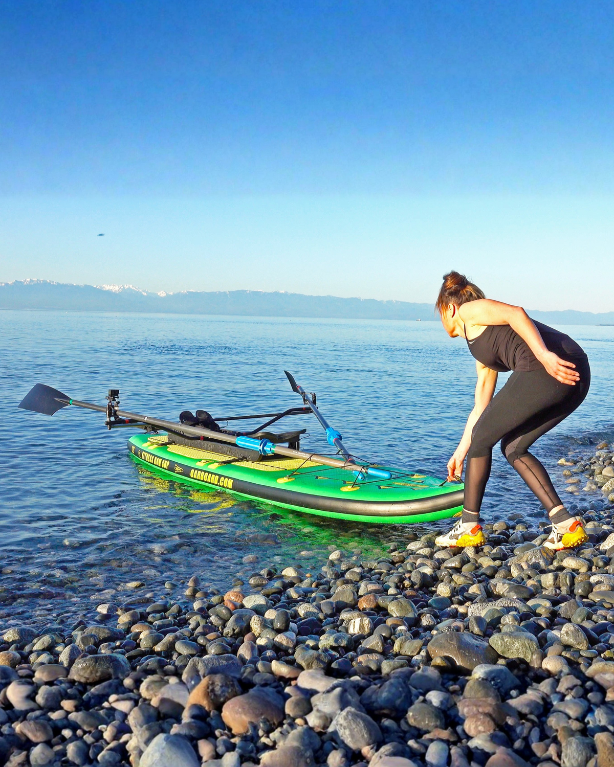 A woman in black activewear bends down next to a green inflatable stand-up paddle board on a rocky shoreline, with calm blue water and distant mountains under a clear sky.