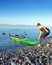 A woman in black activewear bends down next to a green inflatable stand-up paddle board on a rocky shoreline, with calm blue water and distant mountains under a clear sky.