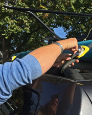 A close-up lifestyle shot of a person's arm and hand securing a black strap to a teal and yellow roof rack mounted on a dark gray car, with trees and blue sky in the background.