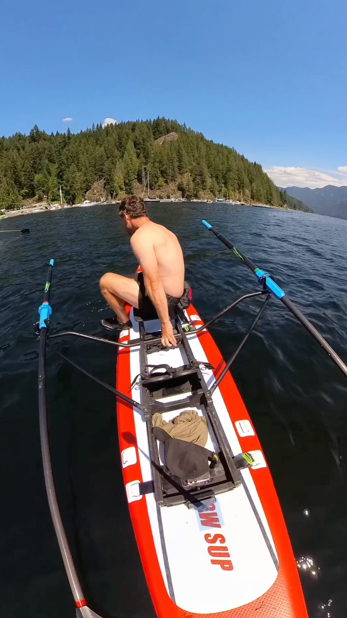 A shirtless man sit on the edge of a red and white paddle board about to jump into the water. Green trees are visible behind him.
