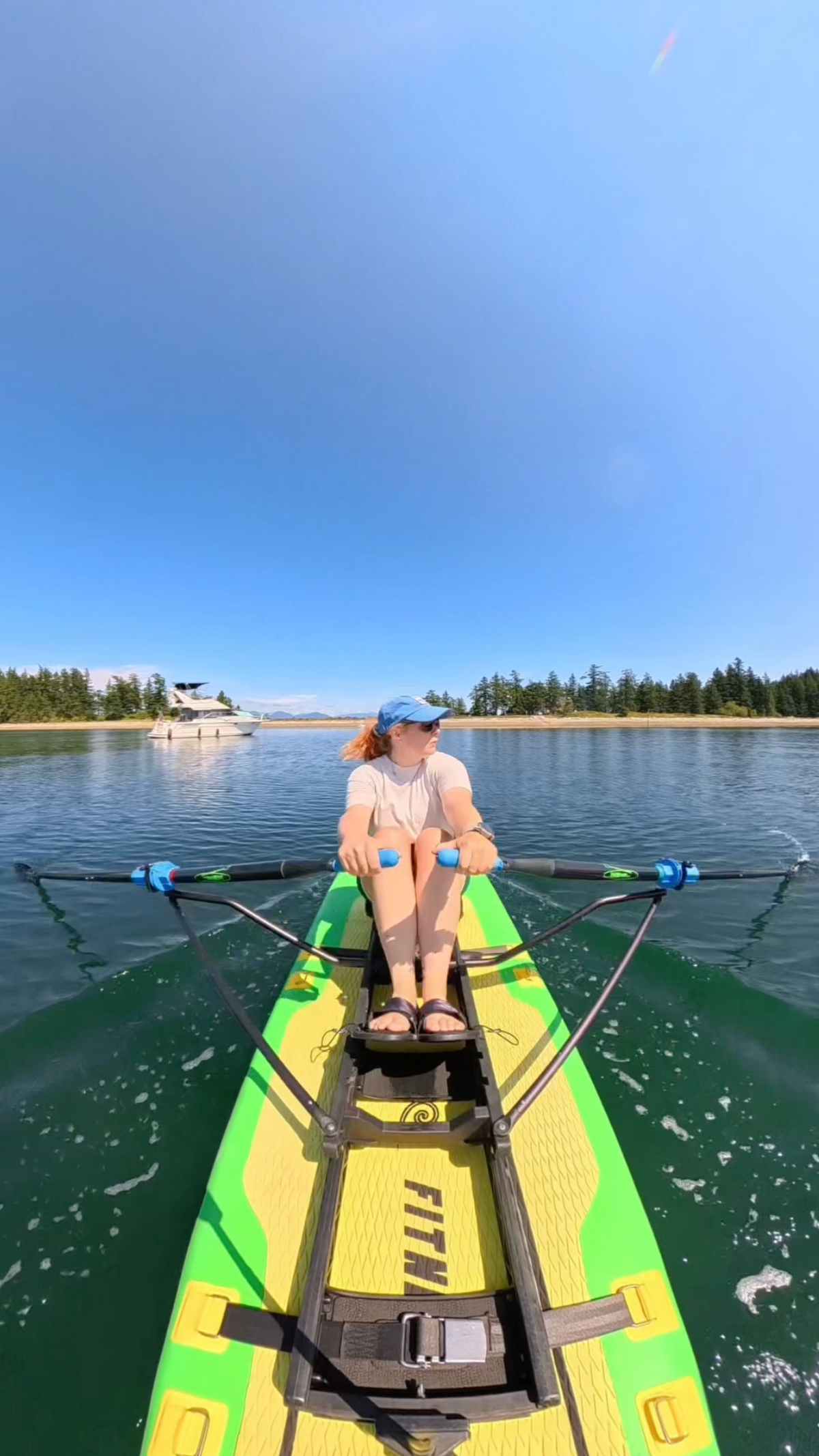 A woman sits on a green and yellow paddleboard on a calm lake, holding the paddle handles. The board has 'FITME' printed on the deck, and trees and boats are visible in the background under a clear blue sky.