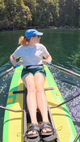 A woman rowing on a bright green and yellow paddleboard on calm green water with a forest shoreline in the background.
