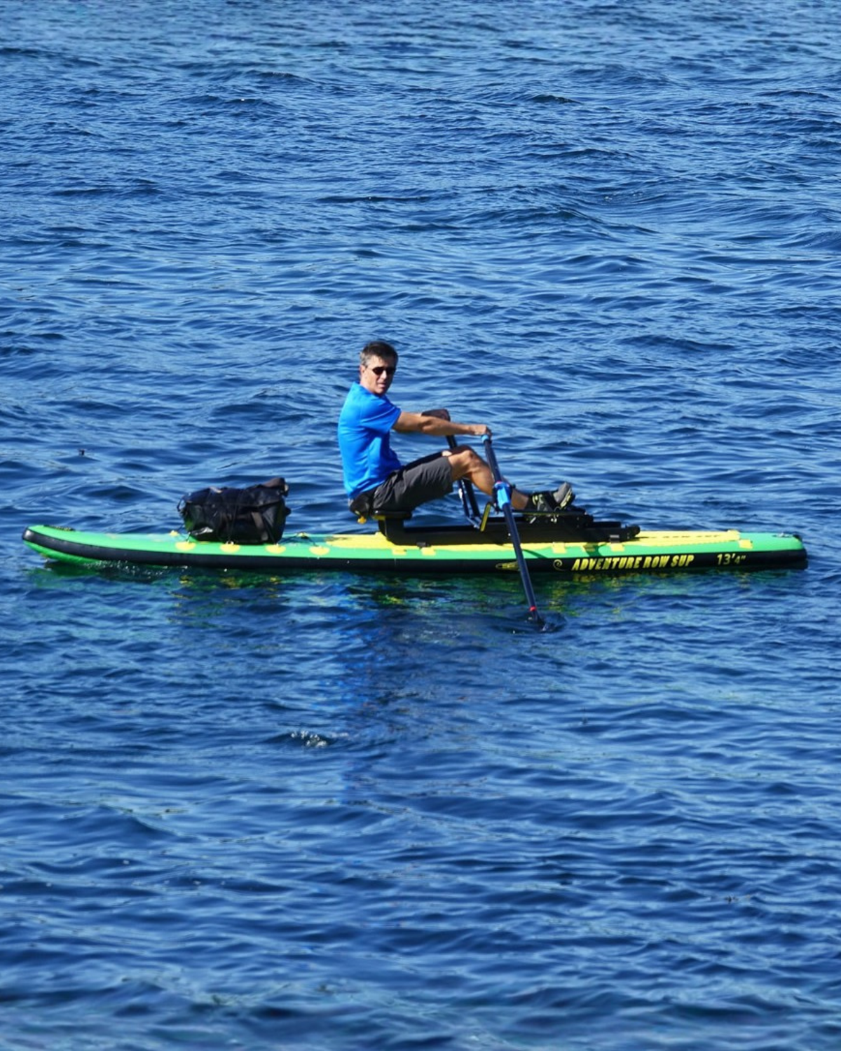 A man in a blue shirt rowing a green paddleboard on blue water, holding oars.  Gear bags are strapped to the front of the board.