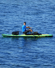 A man in a blue shirt rowing a green paddleboard on blue water, holding oars.  Gear bags are strapped to the front of the board.