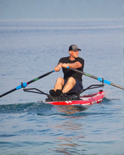 A man rowing a red and white paddle board with a black frame and black oars on blue water.