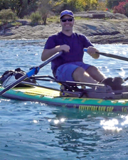 A man wearing a blue cap and sunglasses rows on a paddleboard on blue water, with rocky shoreline and trees in the background. The paddleboard is yellow and green with visible branding text.