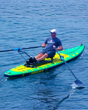 A man rowing on a green and yellow paddleboard with an attached seat frame, photographed on open blue water. The board has visible branding text near the front.