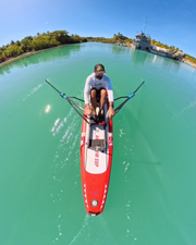 A person rows on a red and white paddleboard in turquoise water, with a tropical shoreline, palm trees, and a boat visible in the background under a clear blue sky.