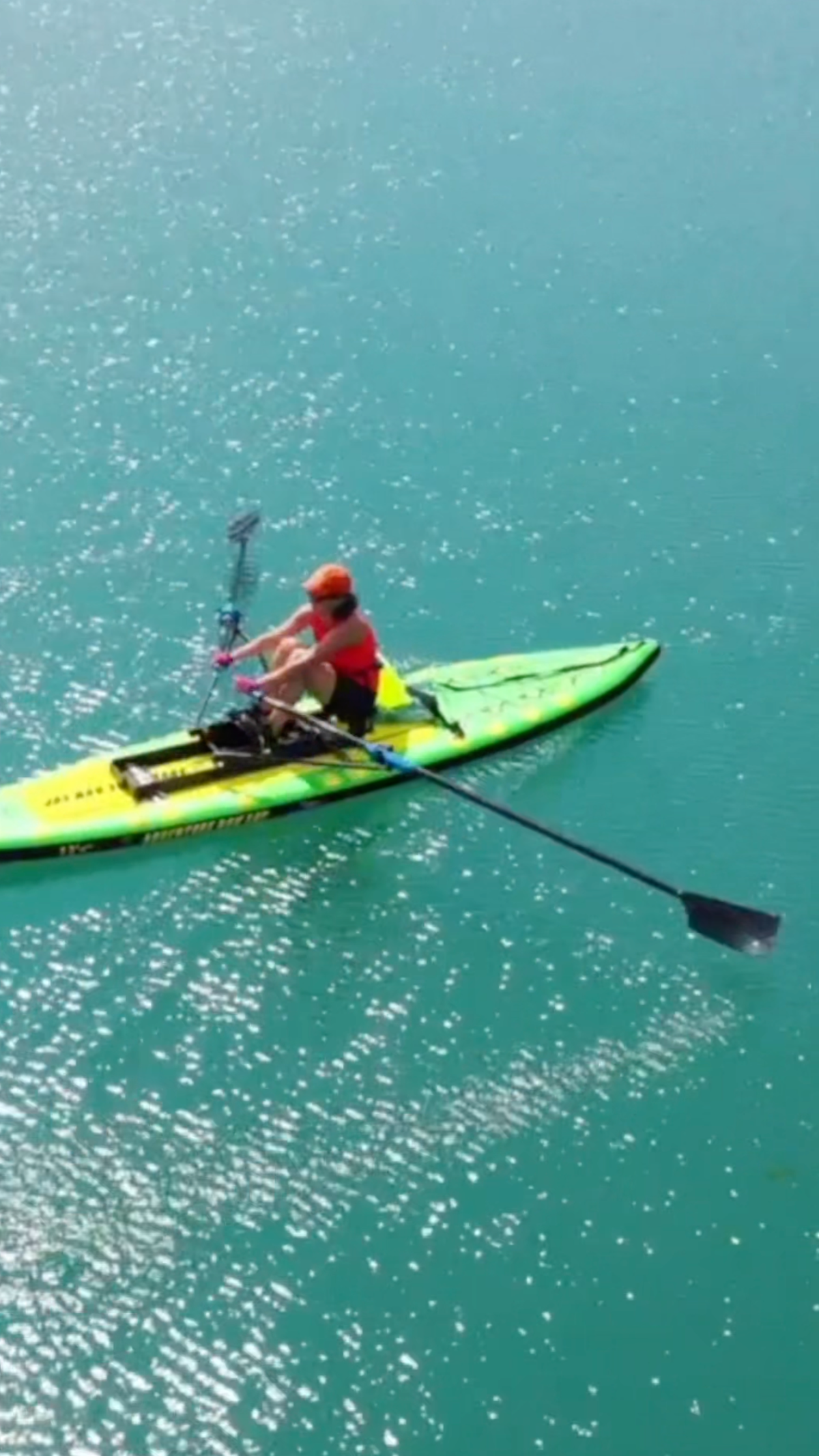 A woman wearing an orange hat and tank top rows a green and yellow paddle board on bright blue water, holding black oars. The photo is taken from above.