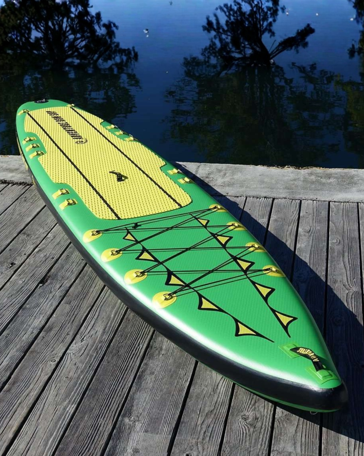 An inflatable stand-up paddle board on a wooden dock with water and trees in the background. The board is green and yellow with black trim and bungee cords.