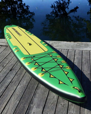 An inflatable stand-up paddle board on a wooden dock with water and trees in the background. The board is green and yellow with black trim and bungee cords.