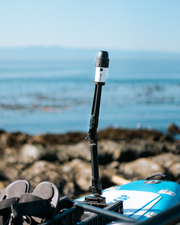 A black and white torch mounted on a pole, set on top of a blue and white paddleboard with a rocky shoreline and ocean in the background.