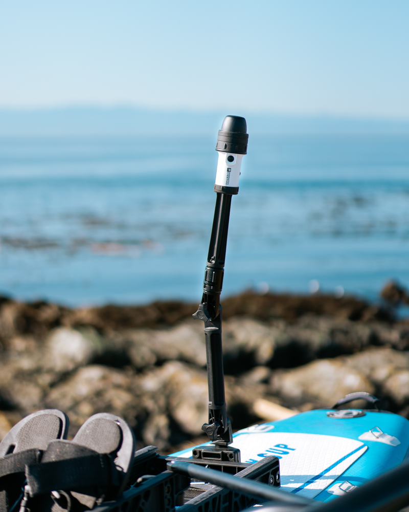 A black and white torch mounted on a pole, set on top of a blue and white paddleboard with a rocky shoreline and ocean in the background.