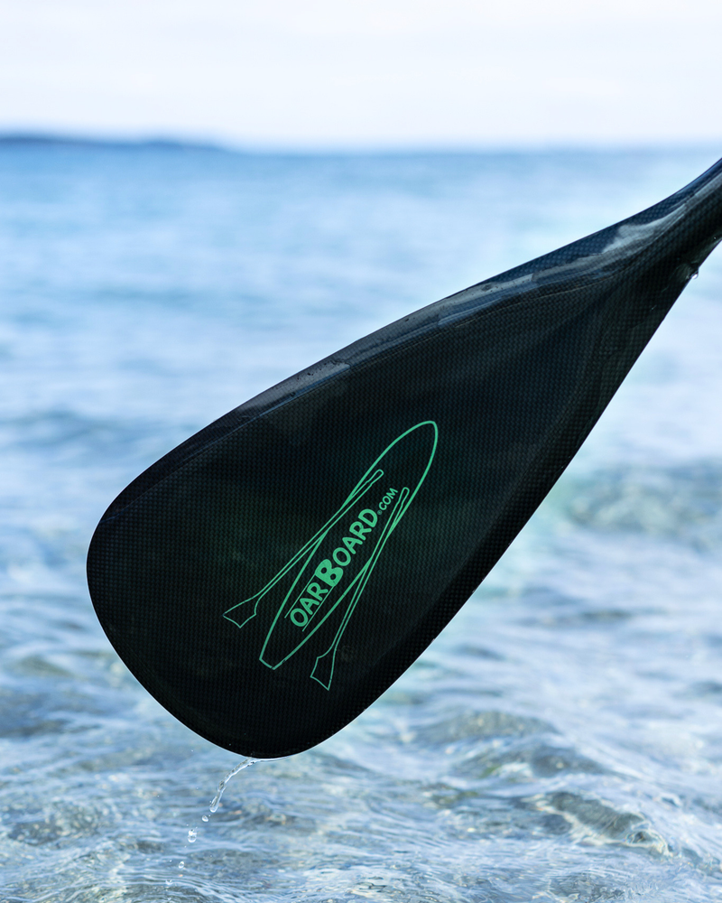 Carbon fibre paddle blade with a green surf board shaped logo with 'OarBoard.com' written on it, photographed on with blurry ocean behind.