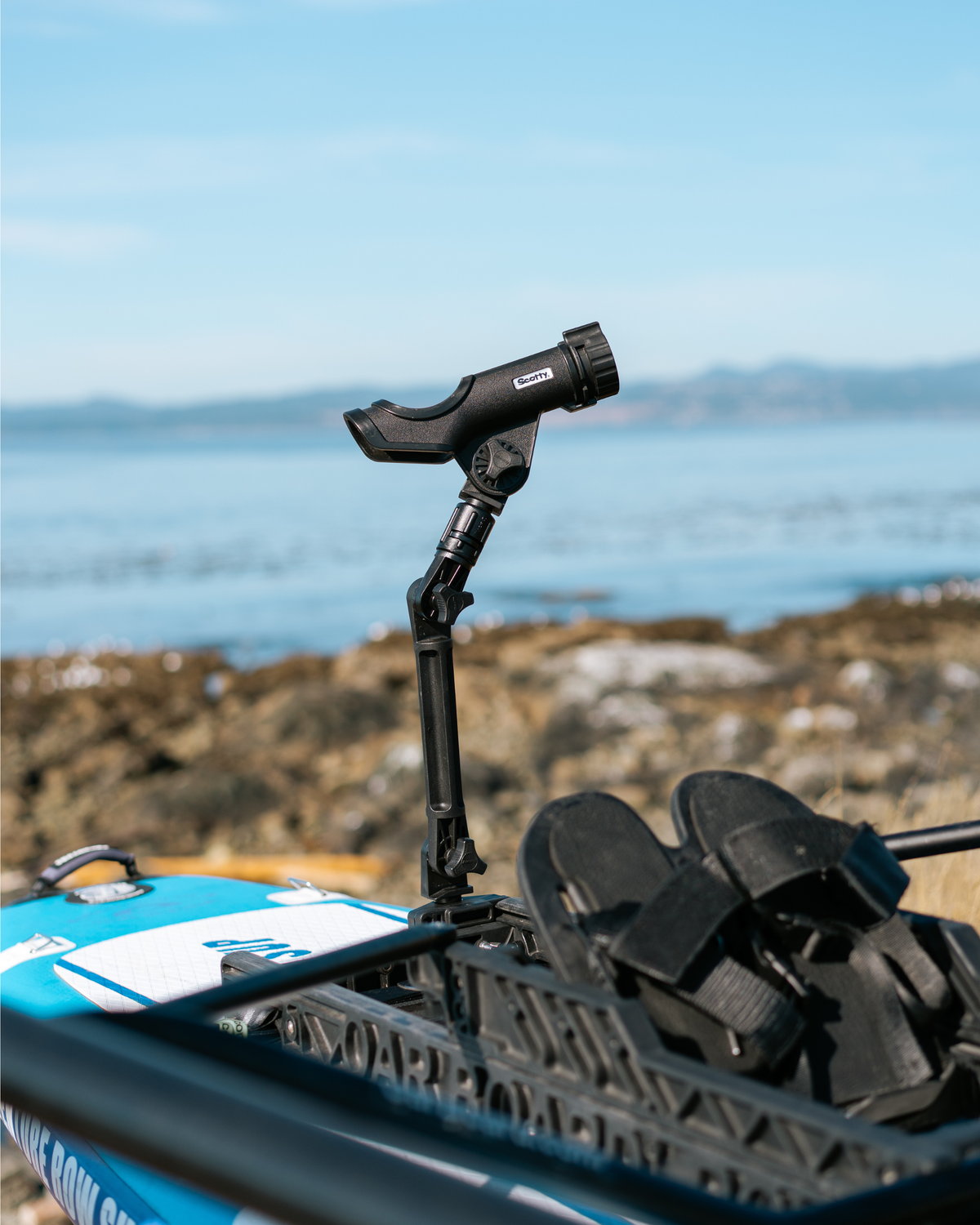A black fishing rod holder, set on a blue and white paddle board with black foot straps, photographed outdoors near a rocky shoreline and water.
