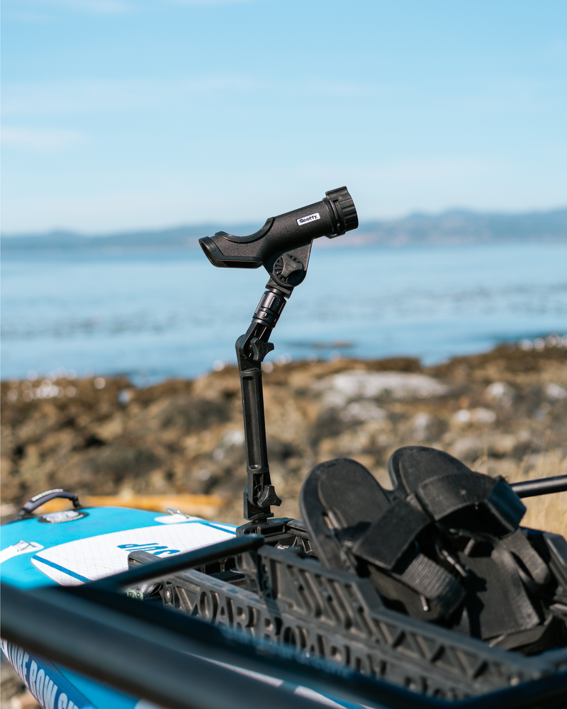A black fishing rod holder, set on a blue and white paddle board with black foot straps, photographed outdoors near a rocky shoreline and water.