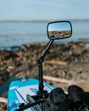 A black adjustable rowboat mirror mounted on a blue and white paddle board, with a rocky shoreline and water blurred in the background.