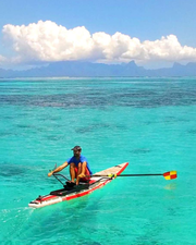 A person rowing a paddle board on bright turquoise ocean water with distant mountains and a large white cloud formation in the sky. The board is red, white, and black, and the person wears a blue shirt and orange shorts.