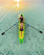 A person rowing on a bright green and yellow stand-up paddle board in clear turquoise water at sunset, viewed from above.