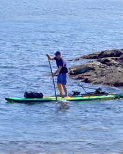 A man paddles a paddleboard on blue water near rocky shoreline, wearing a dark blue shirt, blue shorts, and a dark cap. The board is green and yellow with black gear and accessories.
