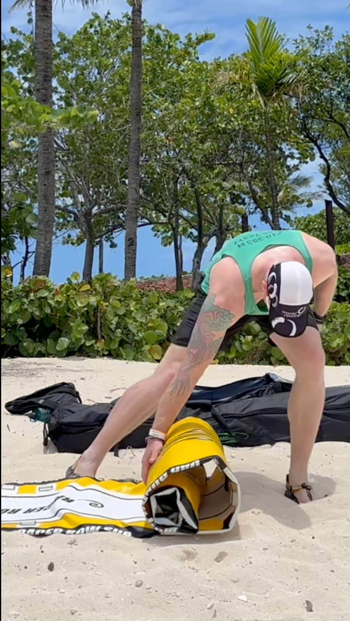A man on a sandy beach bends down to roll out yellow and white paddle board  next to a black bag, with palm trees and blue sky in the background.