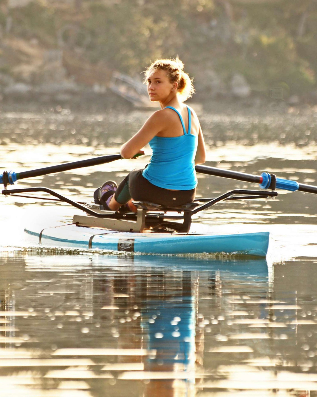 A woman rowing on a calm lake on paddle board wearing a bright blue tank top and black shorts, with warm golden sunlight reflecting on the water.