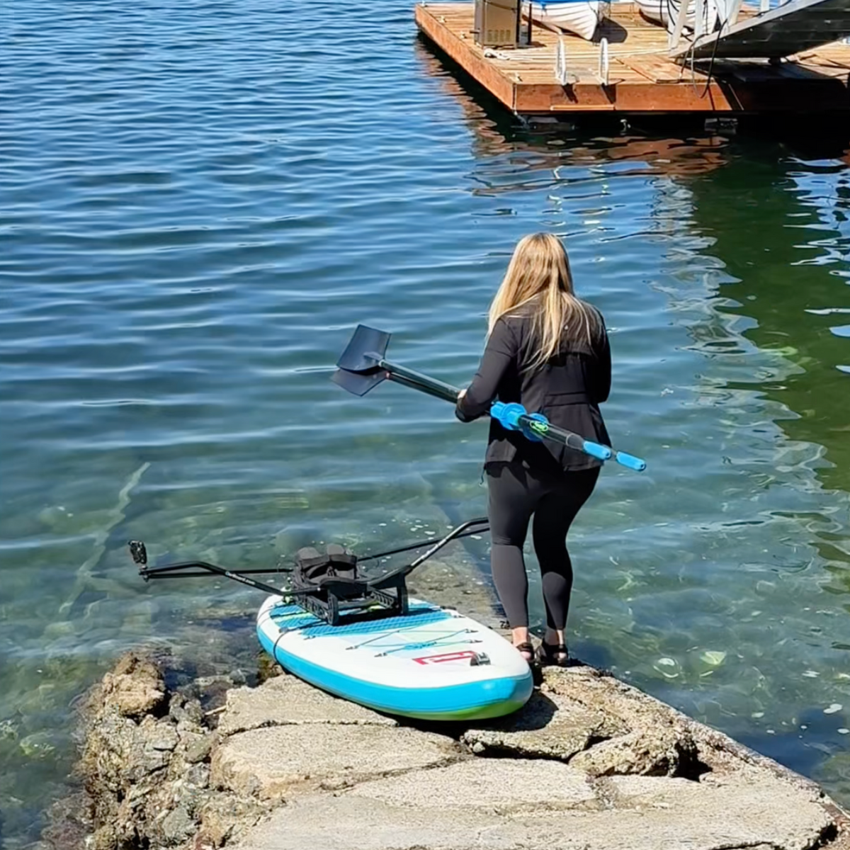 A woman in a black wetsuit stands in shallow water holding a oars next to a paddleboard on rocks by the shoreline, with a wooden dock and blue water in the background.