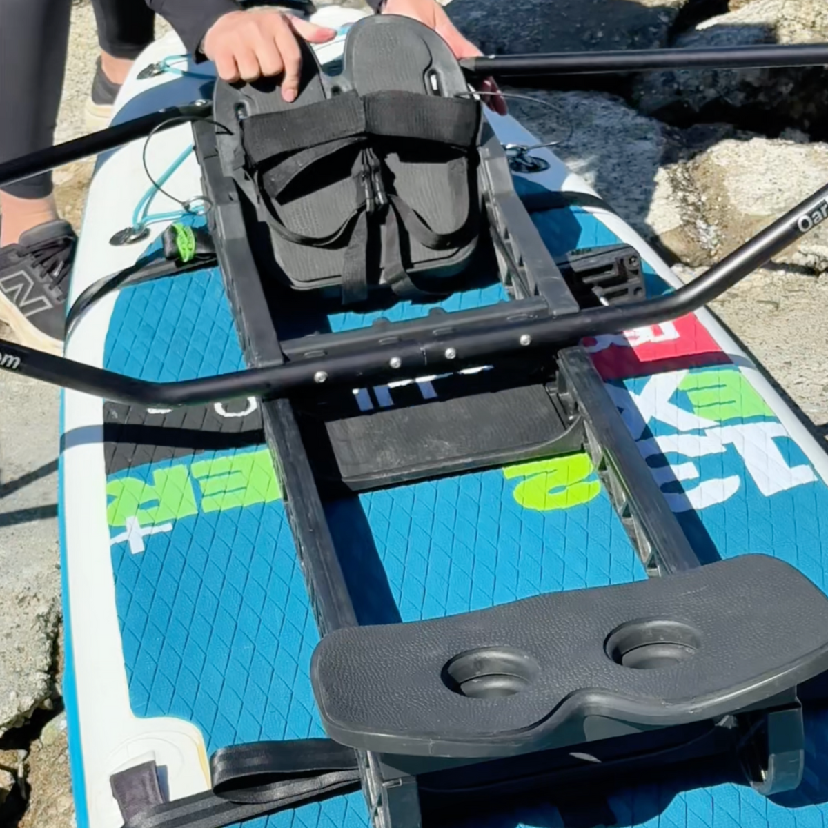 Close-up of a blue and white stand-up paddle board with a black mounting frame, black footrests, and a black dual cup holder, shown outdoors on a rocky surface with part of a person's hand and legs visible.