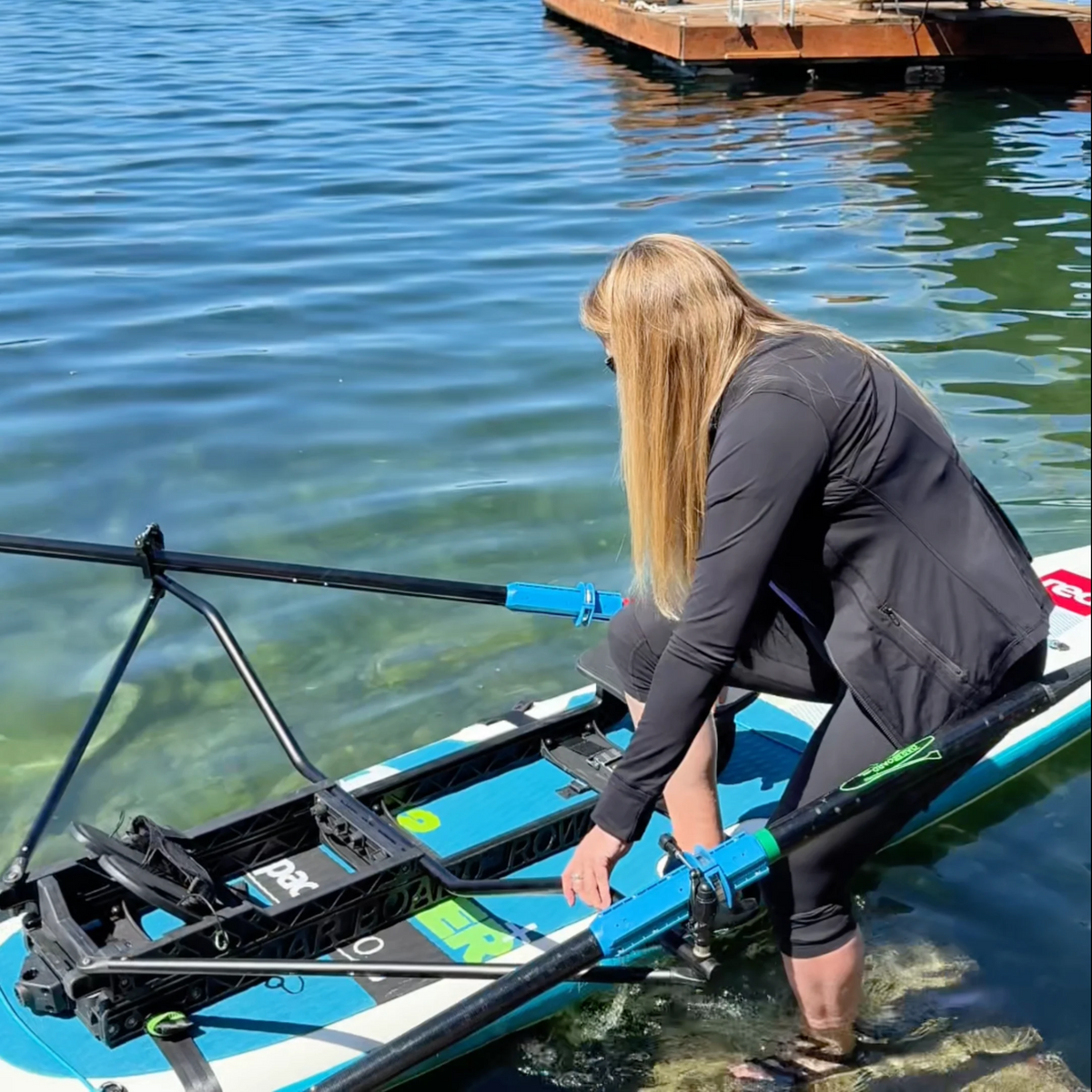 A woman in a black long-sleeve athletic top and black leggings is crouching on a blue and white stand-up paddleboard in shallow blue water, holding a black oar with bright blue and green accents.