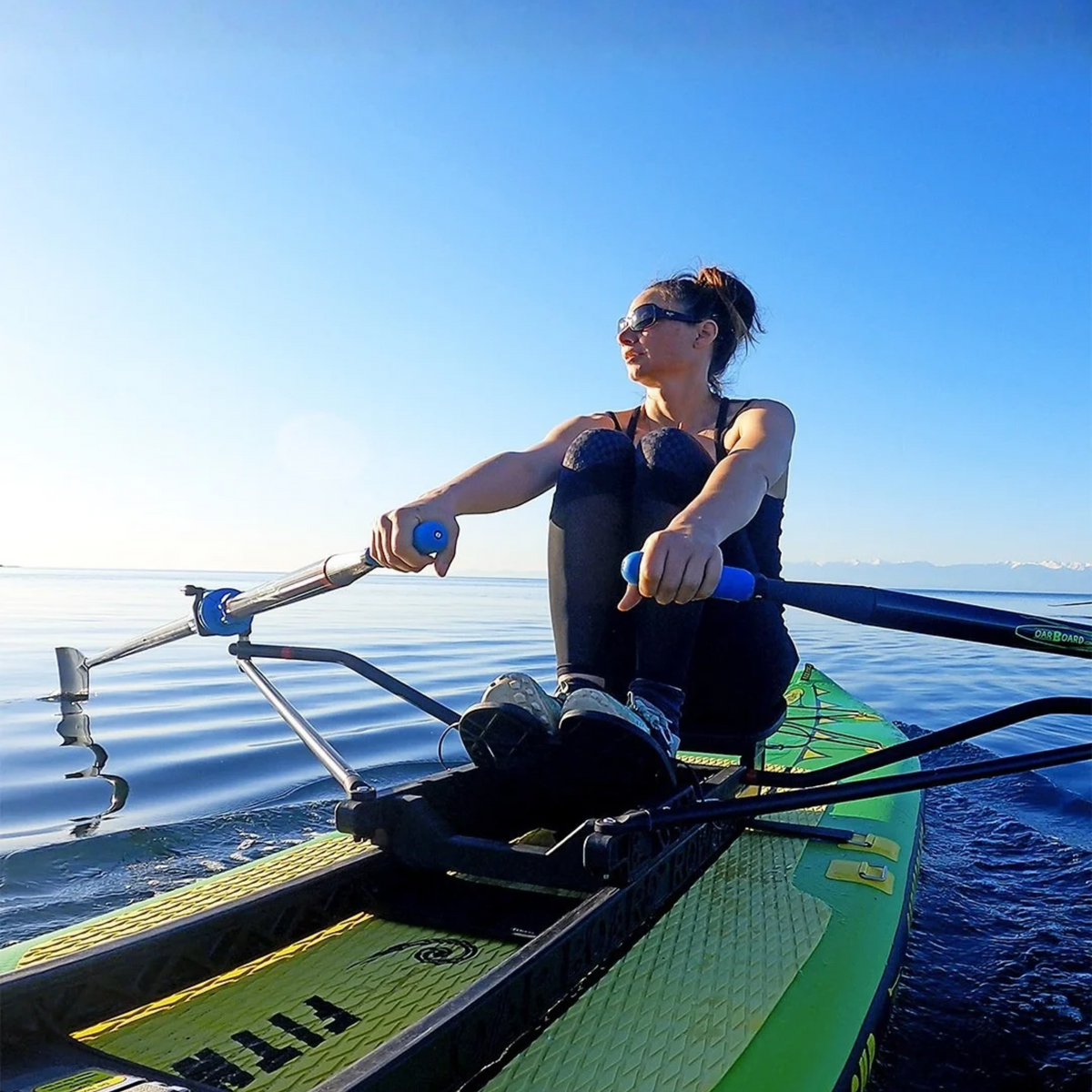 A woman rowing on a green and yellow stand-up paddleboard on calm blue water under a clear blue sky.