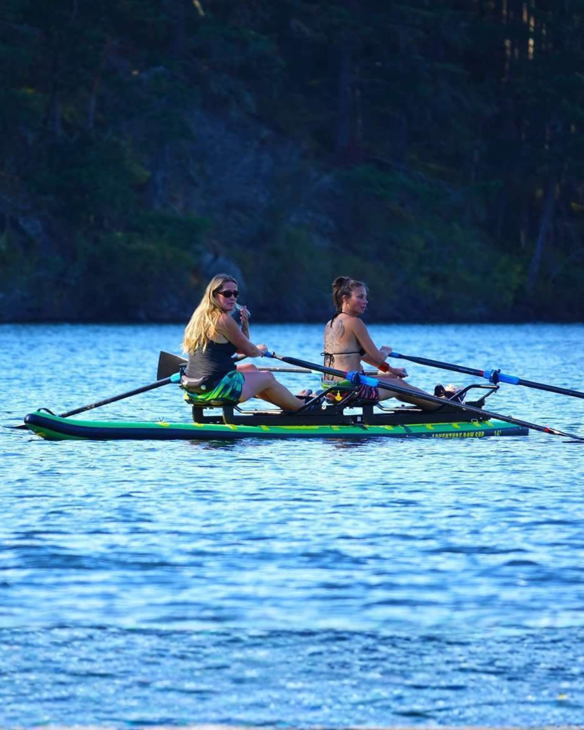 Two women rowing on a tandem paddleboard on a calm blue lake with a dark forest shoreline in the background.