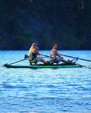 Two women rowing on a tandem paddleboard on a calm blue lake with a dark forest shoreline in the background.