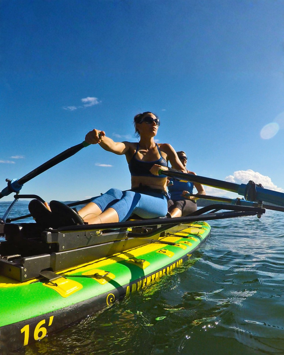 A woman rows a bright green and yellow inflatable paddle board on open water under a clear blue sky, with another person seated behind her.