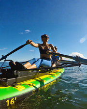 A woman rows a bright green and yellow inflatable paddle board on open water under a clear blue sky, with another person seated behind her.