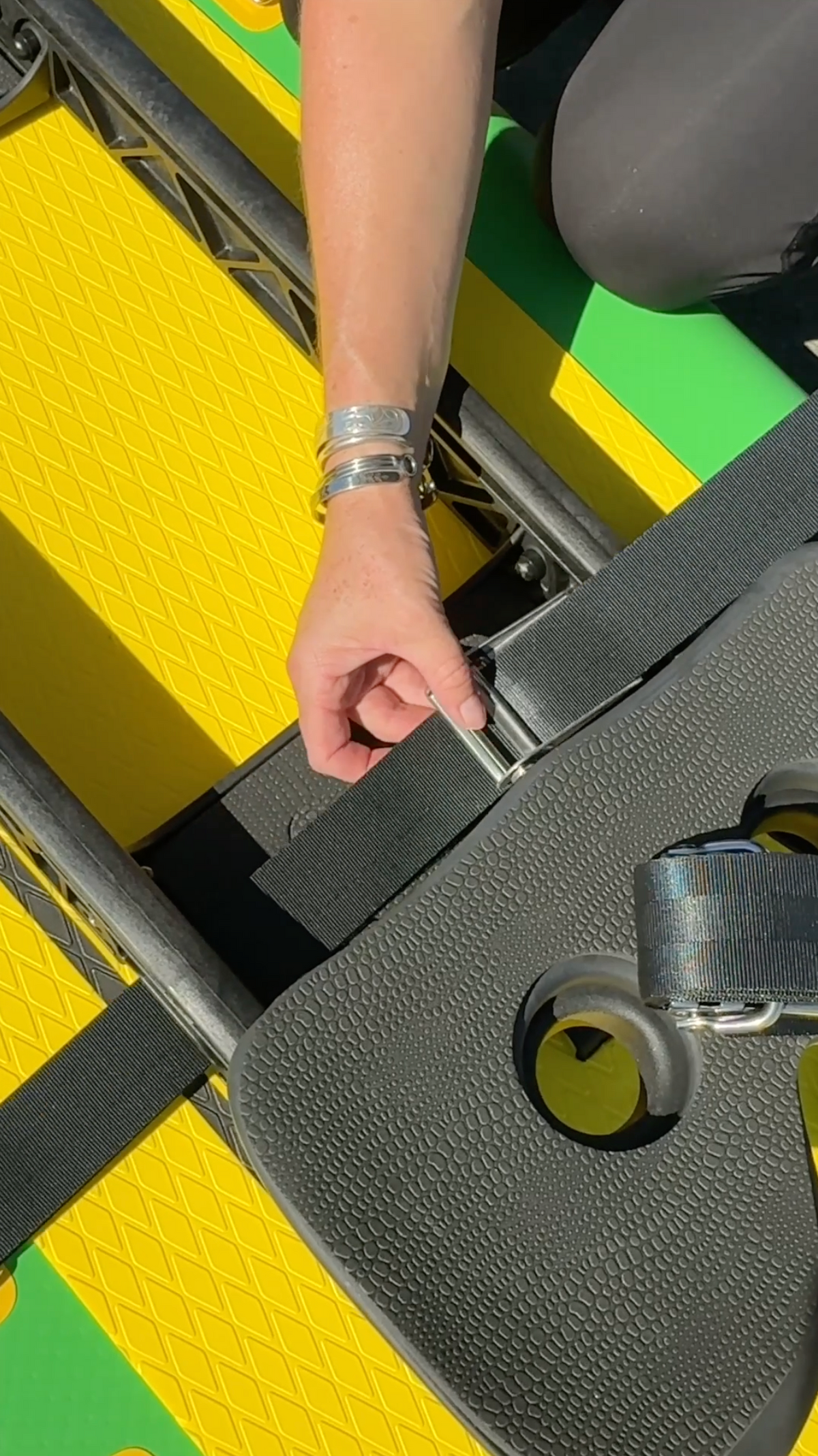 A close-up of a person's hand adjusting a black strap on a yellow and green paddle board in bright sunlight.