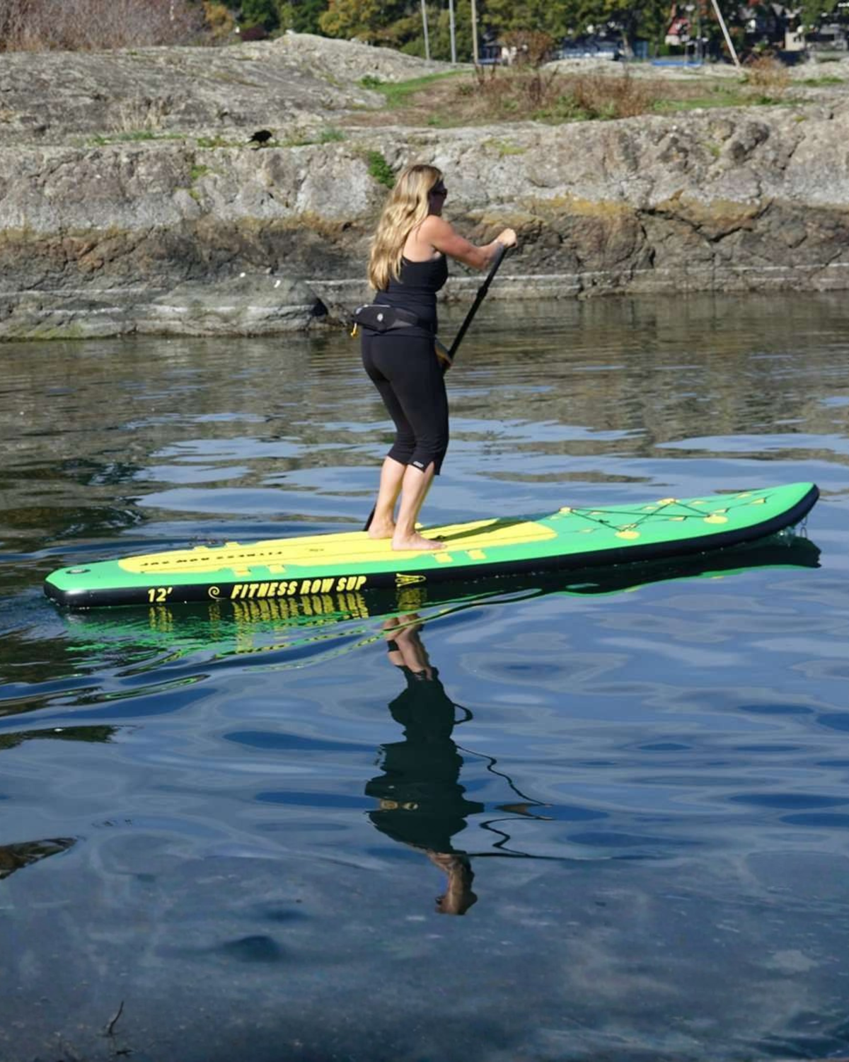 A woman stands on a green and yellow stand-up paddleboard on calm water, holding a paddle. The background shows a rocky shoreline with some trees and a street visible in the distance.