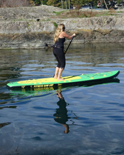 A woman stands on a green and yellow stand-up paddleboard on calm water, holding a paddle. The background shows a rocky shoreline with some trees and a street visible in the distance.