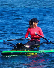 A woman in a red long-sleeve top, black leggings, and a black cap rows on a green and black paddleboard on blue water. The board has yellow text reading 'FITNESS ROW SUP'.