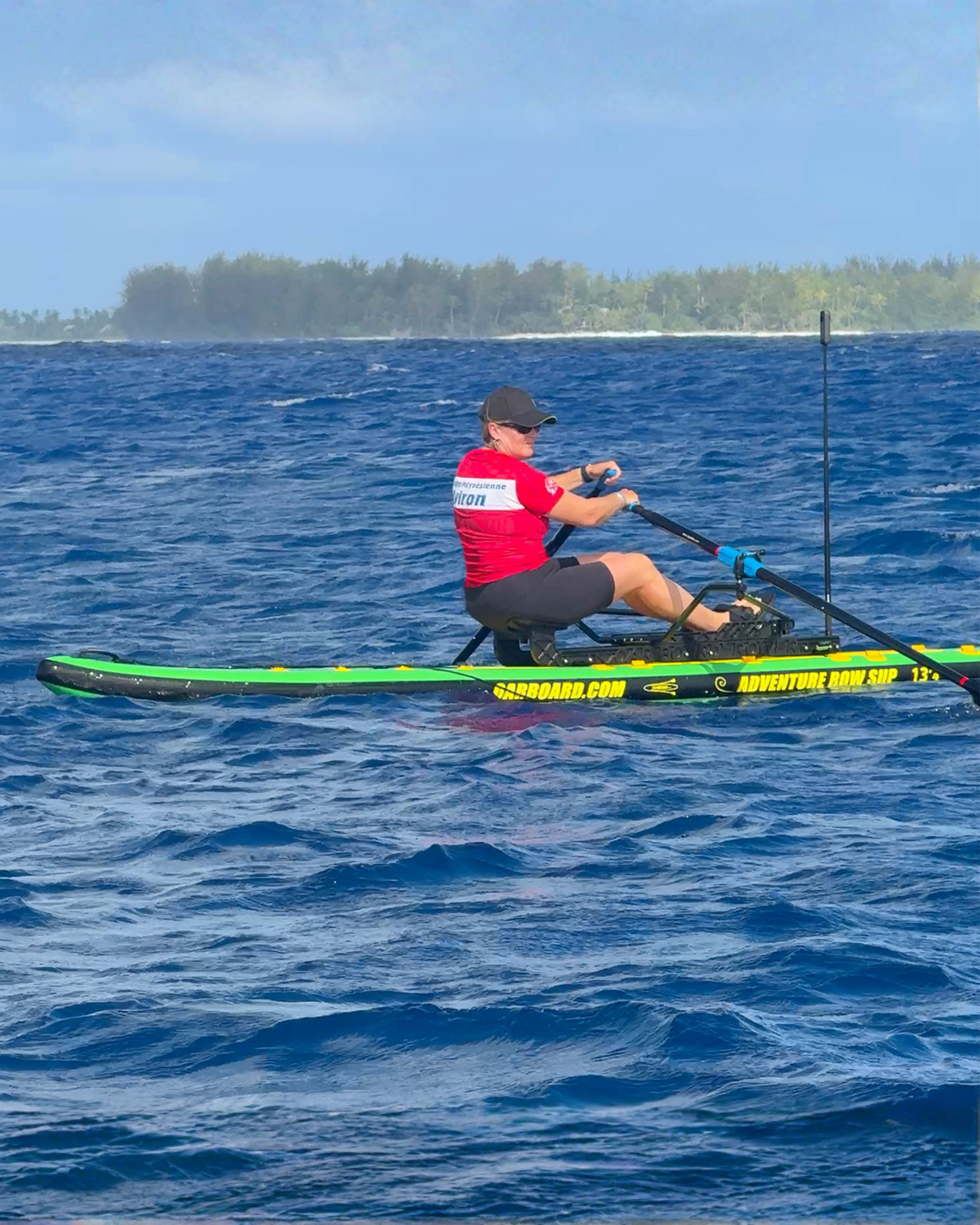A person rowing on a green and black stand-up paddleboard on blue ocean water, with a tree-lined island visible in the background under a blue sky.