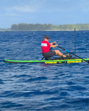 A person rowing on a green and black stand-up paddleboard on blue ocean water, with a tree-lined island visible in the background under a blue sky.