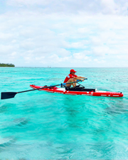 A person in a red cap and red top rows a red and white paddle board on bright turquoise ocean water under a cloudy sky.