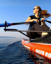 A woman rowing on a red and white paddle board on open water under a clear blue sky, wearing a black t-shirt and black leggings. The board has 'RACER ROW' printed on the side.