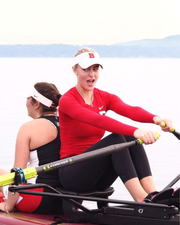 Two women rowing on a lake, with the front rower wearing a red long-sleeve athletic top, black leggings, and a white visor with a red letter 'B'.