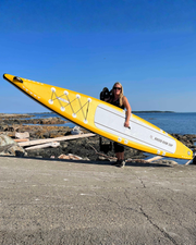 A woman stands on a rocky shoreline holding a large yellow and white stand-up paddleboard on a support frame, with blue sky and water in the background.