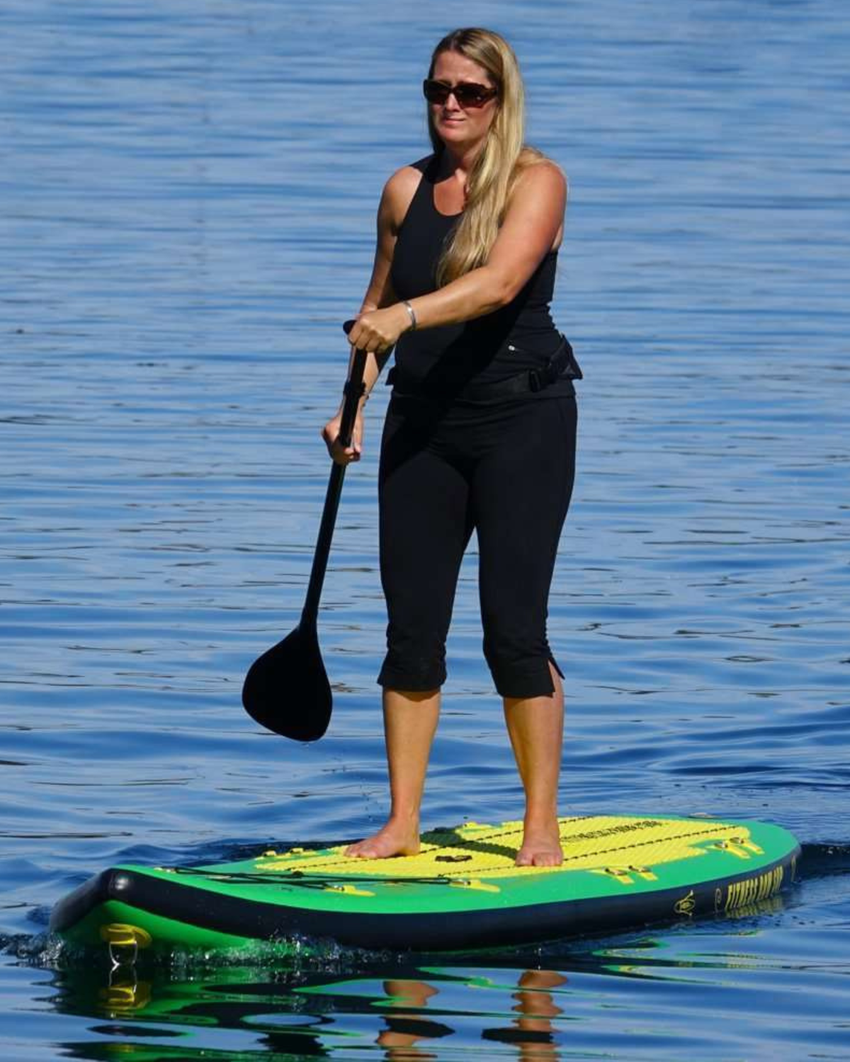 A woman stands on a paddleboard on blue water, holding a black paddle. She is wearing a black tank top, black capri leggings, and sunglasses.