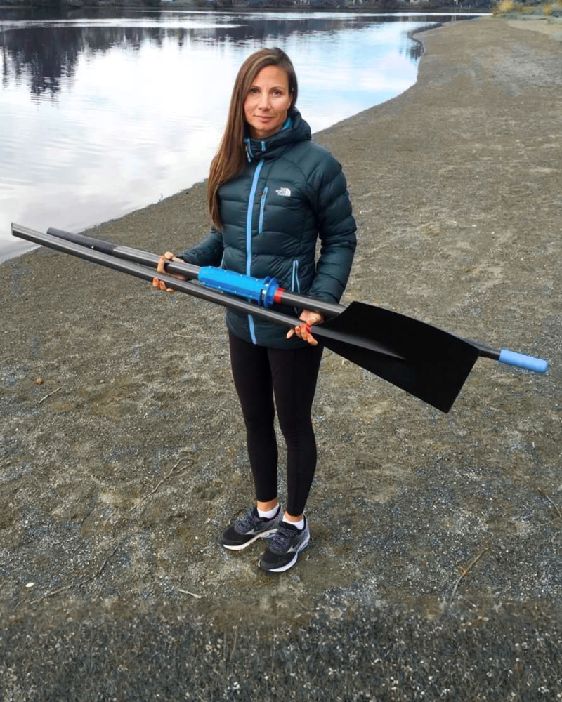 A woman stands on a rocky lakeshore holding a long black rowing blade with blue and gray shaft sections. She is wearing a dark teal puffer jacket, black leggings, and black-and-white running shoes.