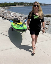 A woman wearing a black 'Rowing' t-shirt and black bike shorts walks on a concrete boardwalk next to the water, pulling a green and yellow paddle board on a dolly.
