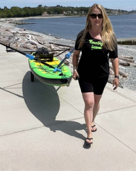 A woman wearing a black 'Rowing' t-shirt and black bike shorts walks on a concrete boardwalk next to the water, pulling a green and yellow paddle board on a dolly.