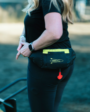 A woman wearing a black pfd lifebelt with a bright yellow-green reflective stripe and a small red hanging toggle, photographed outdoors in a blurred natural setting.